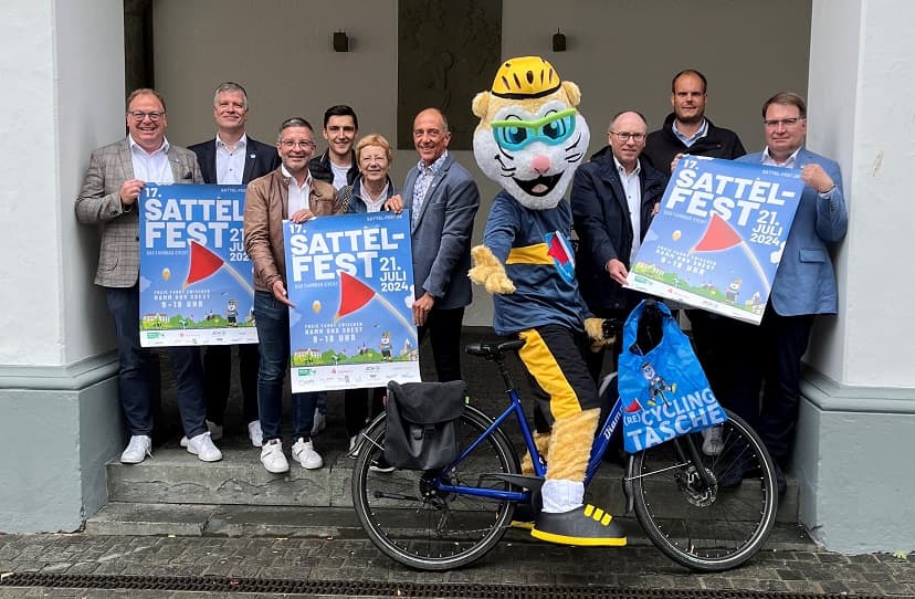 Menschen präsentieren die Plakate zum Sattelfest und das Maskottchen steht mit Fahrrad dabei