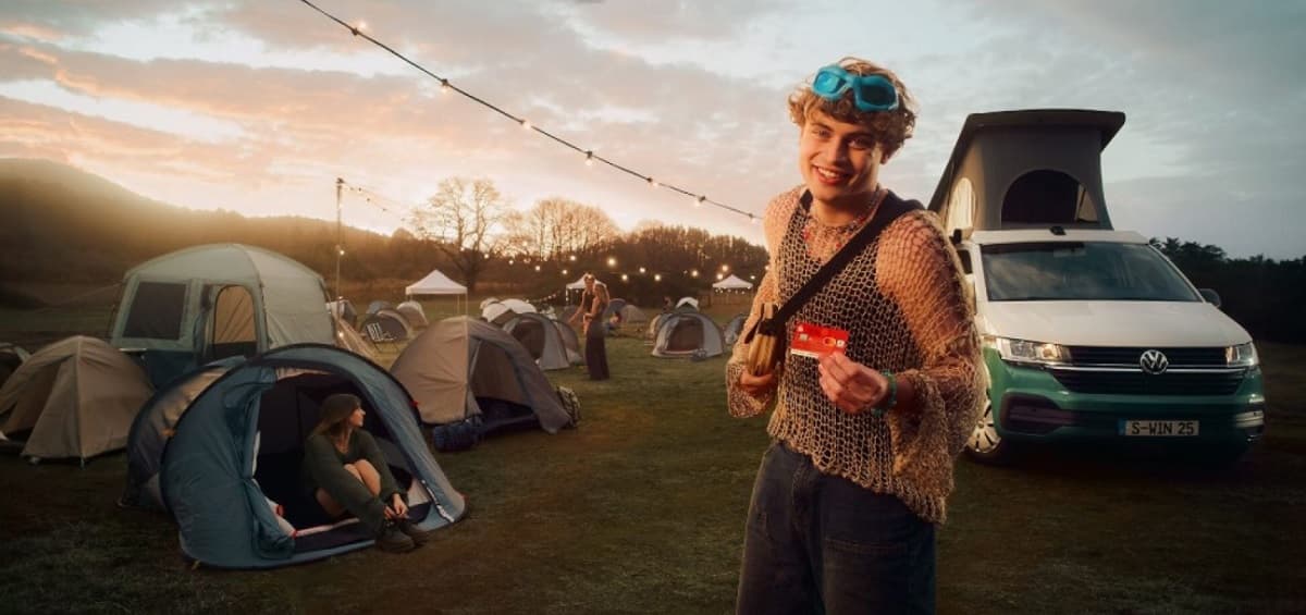 Gewinnspiel Zeltplatz mit einigen Zelten und einem VW Bulli mit aufgestellten Dach und ein junger Mann hält die Sparkassenkarte in die Kamera
