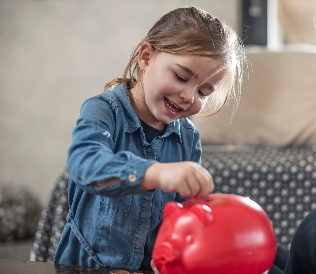 Girl inserting coin from mother into red piggy bank on coffee table